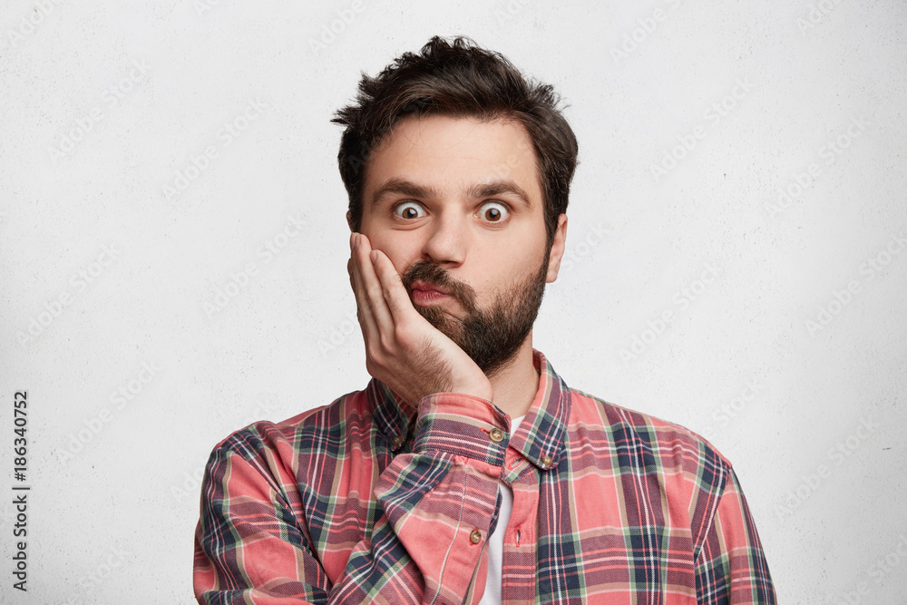 Isolated shot of hesitant shocked male with trendy hairdo, dark beard and mustache, keeps hand on cheek and curves lips, looks in bewilderment and surprisment, isolated over white background.
