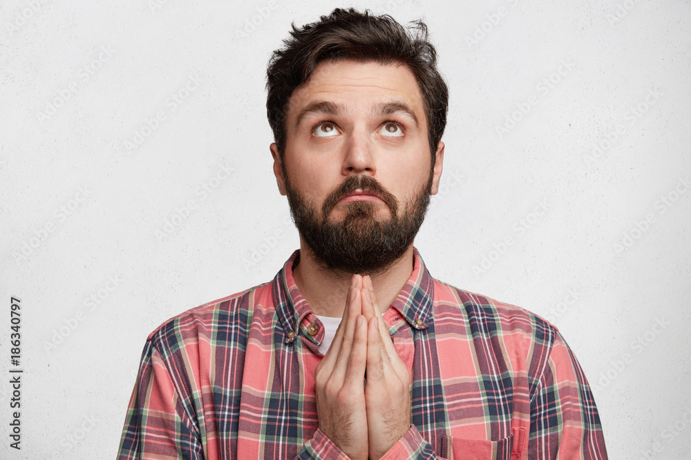 Fototapeta premium Serious bearded male prayer with palms pressed together, looks upwards, dressed casually, prays for good luck or health of relatives, isolated over white background. Religious guy worships success