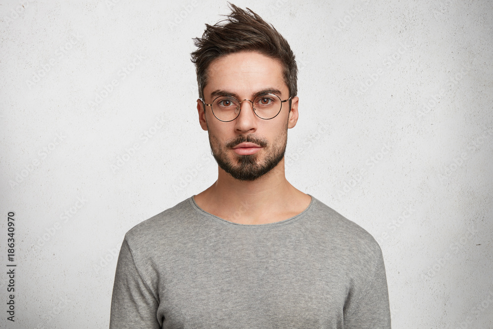 Stylish bearded male student wears round spectacles, has trendy hairstyle, looks confidently, thinks about coming session, isolated over white concrete background. People and human expressions Stylish bearded male student wears round spectacles, has trendy hairstyle, looks confidently, thinks about coming session, isolated over white concrete background. People and human expressions