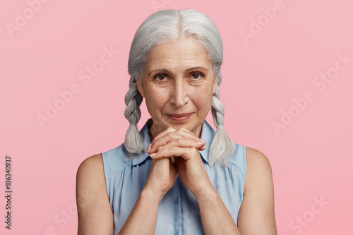 Glad beautiful senior woman looks happily at camera, keeps hands together, being pleased to hear positive news, isolated over pink studio background. Smiling delightful female with grey hair photo