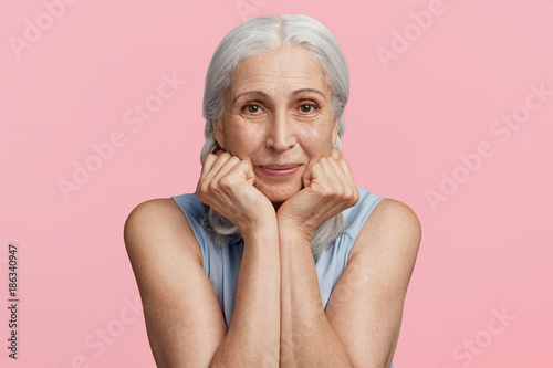 Portrait of positive pleasant looking mature woman with grey hair keeps hands under chin, looks happily directly into camera, expresses positive emotions, isolated over pink background in studio photo