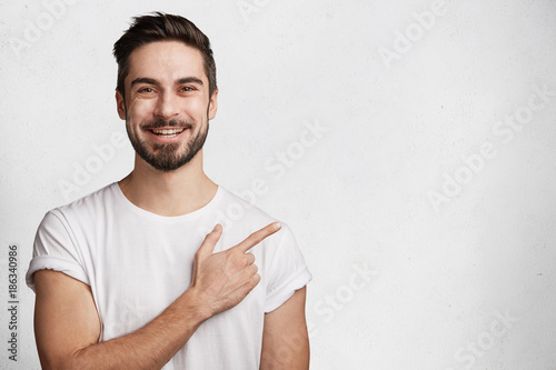 Horizontal portrait of bearded cheerful man has smile, wears casual white t shirt, indicates with fore finger at copy space for your promotional text or advertisment, isolated over concrete wall