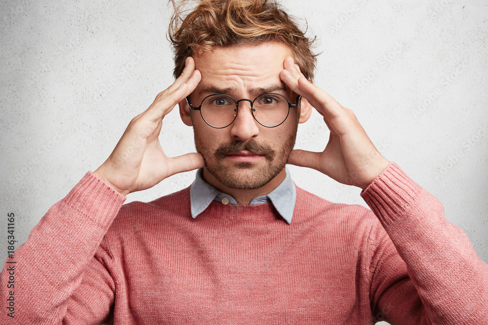 Concentated handsome young unshaven male wears big glasses and sweater, keeps hands on temples as tries to focuse at something, has serious expression, poses against white concrete background Concentated handsome young unshaven male wears big glasses and sweater, keeps hands on temples as tries to focuse at something, has serious expression, poses against white concrete background