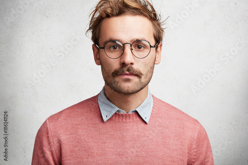 Isolated shot of confident bearded male wears round spectacles and sweater, looks confidently into camera, listens to important information, poses against white concrete background. Facial expressions