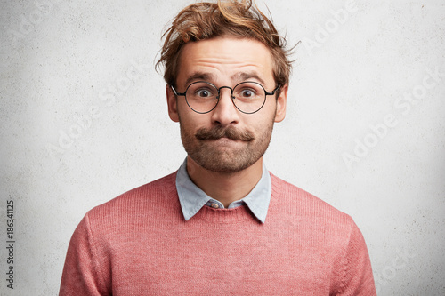 Clumsy male geek presses lips, looks in bewilderment, feels embarrassed in presence of beautiful woman, wears round big spectacles, isolated over white concrete background. Excited emotional male