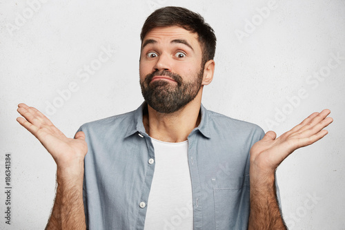 Maybe yes or no. Doubtful bearded fashionable guy shrugs shoulders in bewilderment, tries to make decision, puzzled what he wants, isolated over white concrete wall. Hesitation and uncertainty