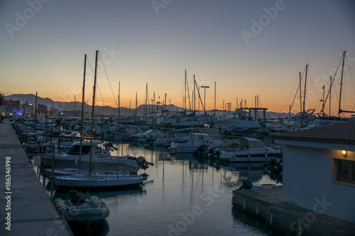 Sunset view of a marina with sailing and motor boats and a mountainous landscape in the background