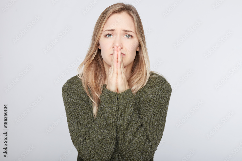 Pleading blond female student rounds lips and has sorrowful expression asks professor for permission to repass exam, wears green warm sweater, isolated over white concrete background studio wall. Pleading blond female student rounds lips and has sorrowful expression asks professor for permission to repass exam, wears green warm sweater, isolated over white concrete background studio wall.