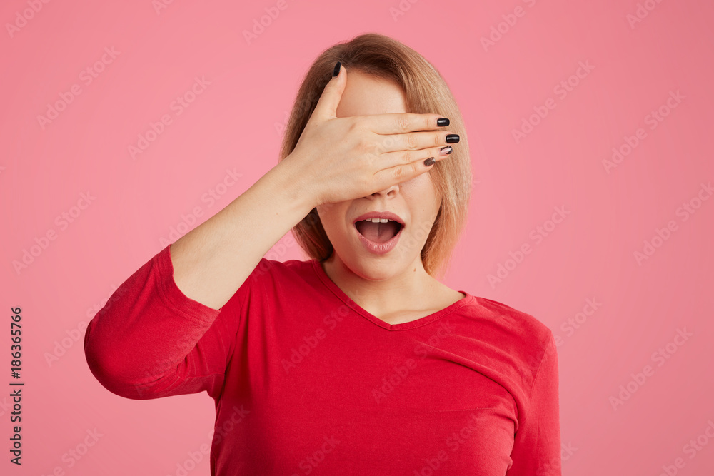Pretty female covers her face with hand, opens mouth, tries to hide from someone, dressed in casual clothes, isolated over pink background. European woman being shocked, covers eyes waits for surprise Pretty female covers her face with hand, opens mouth, tries to hide from someone, dressed in casual clothes, isolated over pink background. European woman being shocked, covers eyes waits for surprise