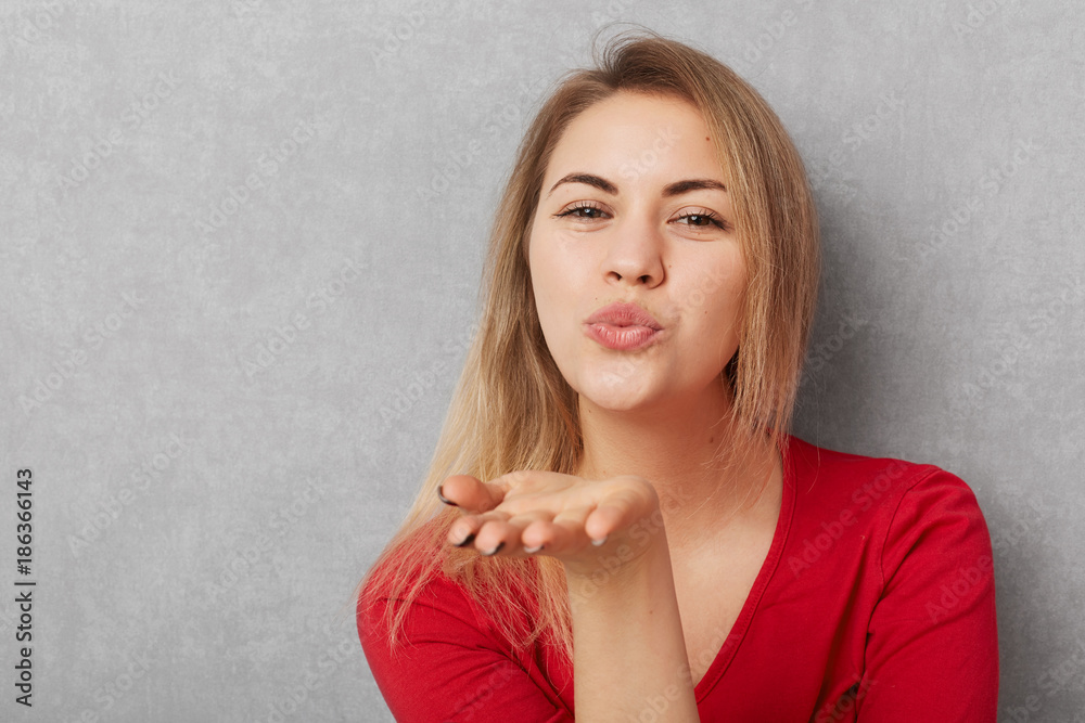 Horizontal shot of beautiful young woman with appealing appearance, makes air kiss, blows at camera, wears red sweater, isolated over grey bckground with copy space for your promotional text