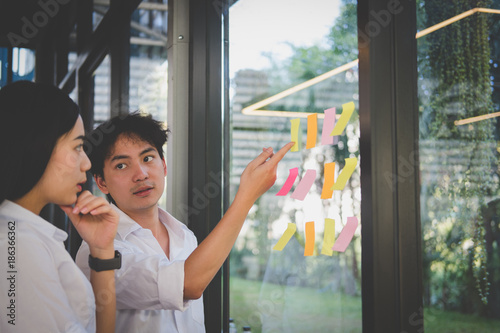 man & woman discussing creative idea with adhesive notes on glass wall at workplace. Sticky note paper reminder schedule at office. business, brainstorming, creativity concept