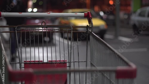 Buyers pass by a shopping trolley in a parking lot near the hypermarket at night. Close-up. The dark background