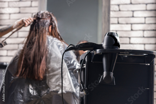 Focus on hair dryer on a special stand near female stylist dyeing hair of young woman