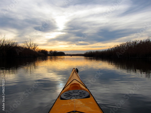 Kayaking on peaceful calm lake or river near dry bulrush at sunset at cloudy winter day. Shot from point of view of the paddler. Bow of yellow kayak