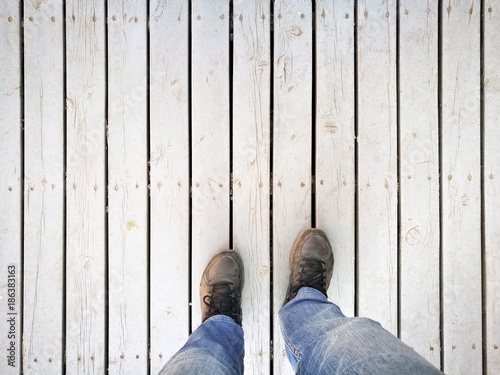 Human feet and legs seen from above. Standing on dirty white wooden pathway; wearing dusty sport shoes and blue jeans in backpacker style - Copy-space for add text is on top.