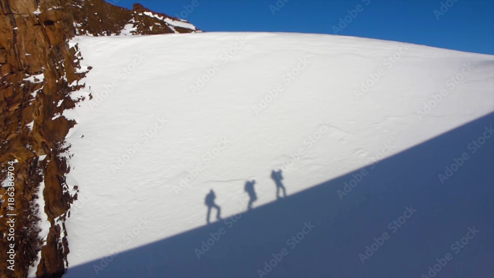 Fototapeta premium The person's shadow on a vertical cliff face. Silhouette of people climbing on overhanging cliff high above clouds and mountains, sun, beautiful colorful sky and clouds behind