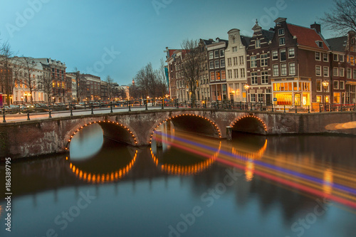 Iconic Amsterdam view with its canals and illuminated boats sailing across and under the narrow bridges pathway, Netherlands
