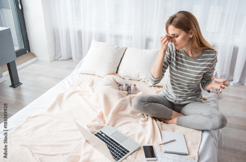 Exhausted young mom resting on bedding. She is holding glass of water in one hand and touching her forehead with another Exhausted young mom resting on bedding. She is holding glass of water in one hand and touching her forehead with another