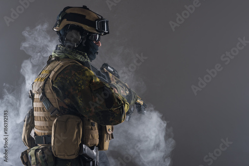 Side view orderly soldier in balaclava and helmet keeping assault carbine in arms. He standing in smoke. Army concept. Copy space