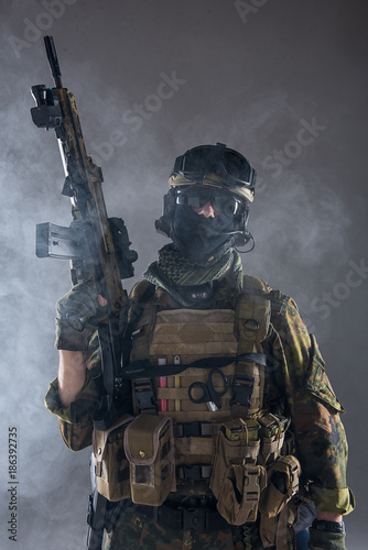 Portrait of serious soldier wearing army clothes while keeping assault rifle in arms. He locating in smoke. Military concept