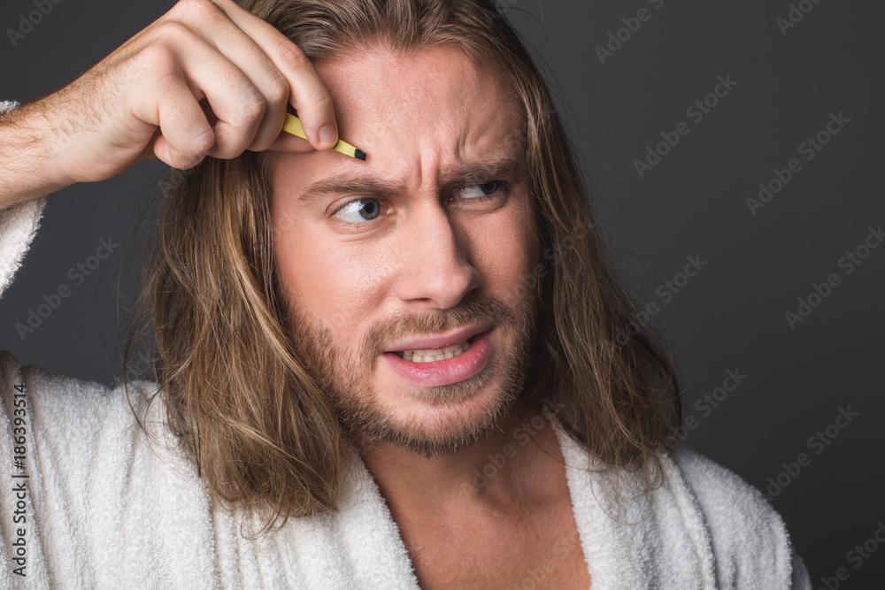 Portrait of displeased man plucking eyebrows, his forehead wrinkled in pain. Isolated on grey background