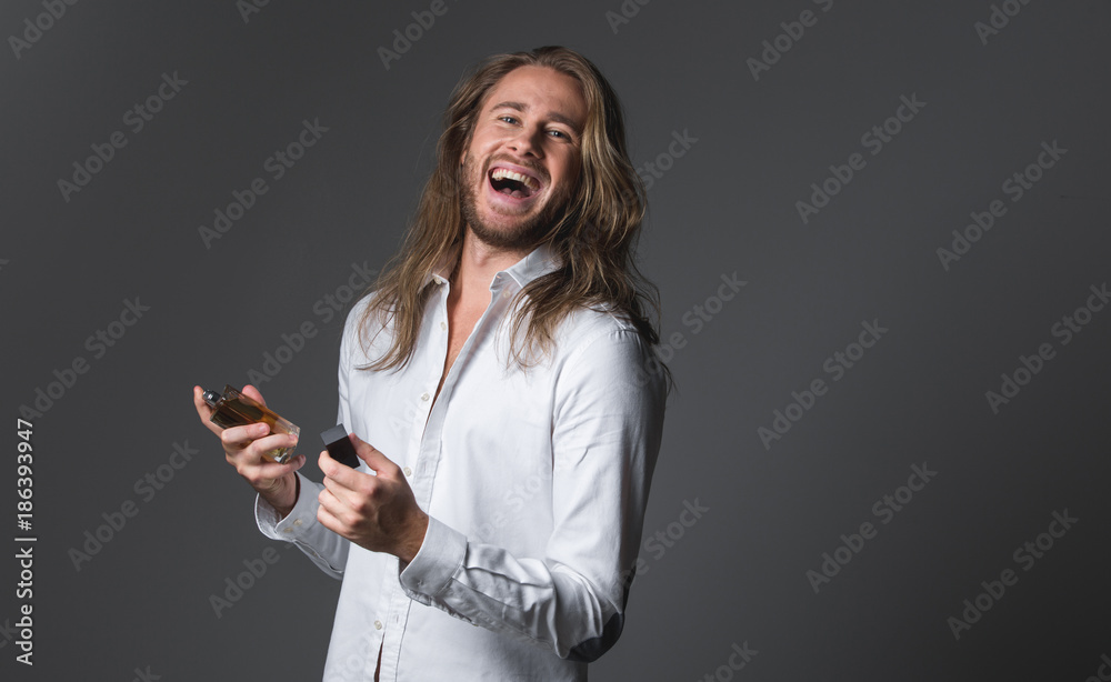 Waist up portrait of cheerful longhaired man holding bottle of essence. He is looking at camera and laughing. Copy space in right side. Isolated on grey background Waist up portrait of cheerful longhaired man holding bottle of essence. He is looking at camera and laughing. Copy space in right side. Isolated on grey background