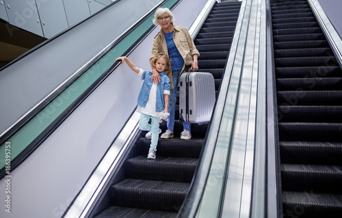 Low angle full length portrait of cheerful elegant senior grandmother is standing on escalator at international airport with her granddaughter while hugging her and holding suitcase. Copy space
