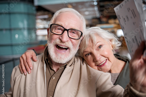 Happy journey. Portrait of delighted old romantic couple is standing at modern terminal. They are looking at camera with joy. Old man is holding his tickets while senior woman is hugging him