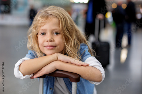 Friendly smile. Portrait of positive charming little girl is leaning on handle of suitcase at international airport. She is waiting for flight while looking at camera with joy. People in background