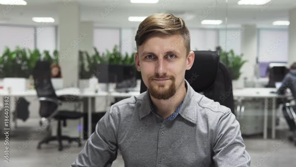 Young business man with a beard smiling at the workplace, office interior as background Young business man with a beard smiling at the workplace, office interior as background
