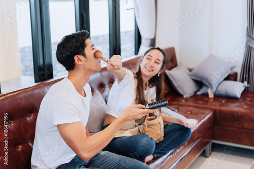 Happy couple funny girlfriend makeup boyfriend watching television comedy show sitting on sofa comfortable couch in living room at home