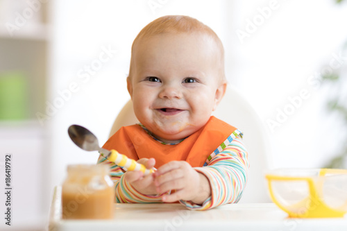 Cheerful baby child eats food itself with spoon. Portrait of happy kid boy in high-chair.