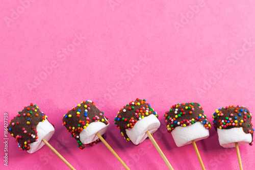 marshmallows on a sticks, laying on pink background. glazed with chocolate and colorful sprinkles.