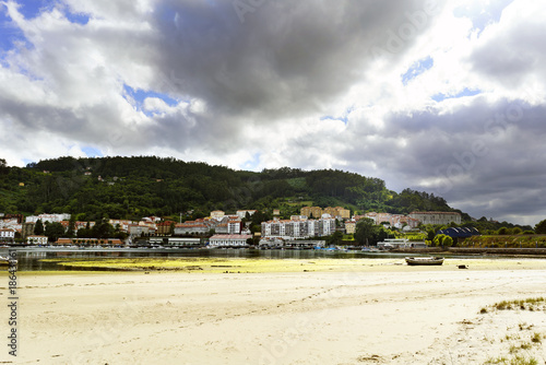 Beach and town of Pontedeume in La Coruna, Spain on the other side of its bay. Sky with clouds threatening rain and lots of vegetation next to the town. A small boat stranded in the sand © peizais
