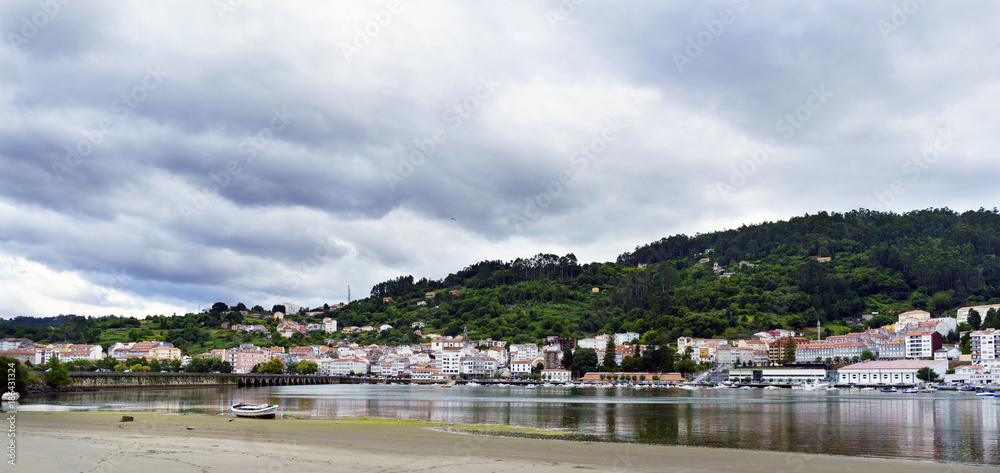 View of the Eume estuary with a fine sand beach in the foreground and a boat stranded in it. On the other side of the river the small town of pontedeume in Galicia, Spain. Sky covered with clouds