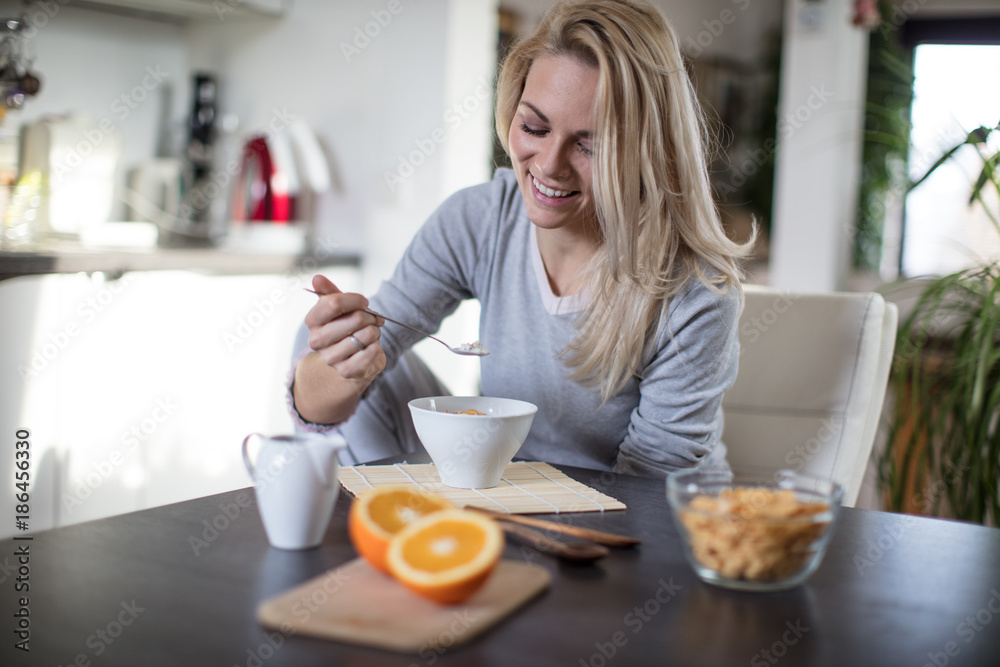Beautiful blond  caucasian woman posing in her kitchen, while drinking coffee or tea and eating a healthy breakfast meal full of cereal and other healthy foods, including fruit