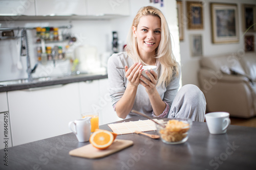 Beautiful blond caucasian woman posing in her kitchen, while drinking coffee or tea and eating a healthy breakfast meal full of cereal and other healthy foods, including fruit