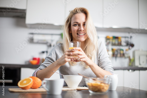 Beautiful blond  caucasian woman posing in her kitchen  while drinking coffee or tea and eating a healthy breakfast meal full of cereal and other healthy foods  including fruit