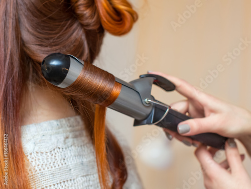 Hairdresser makes hairstyle girl with long red hair in a beauty salon. Create curls with curling irons. Professional hair care.