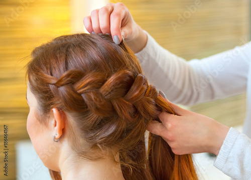 Beautiful girl with long red hair, hairdresser weaves a braid, in a beauty salon. Professional hair care and creating hairstyles.