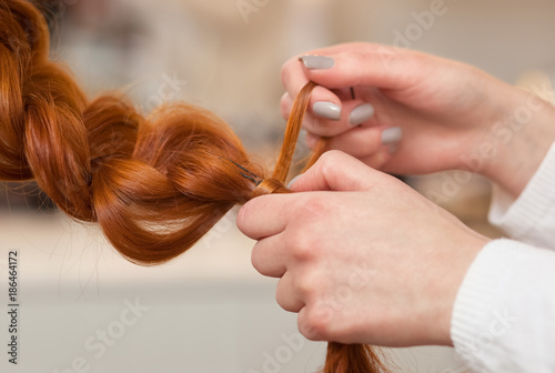 Beautiful girl with long, red-hair, hairdresser weaves a  braid close-up, in a beauty salon. Professional hair care and creating hairstyles.