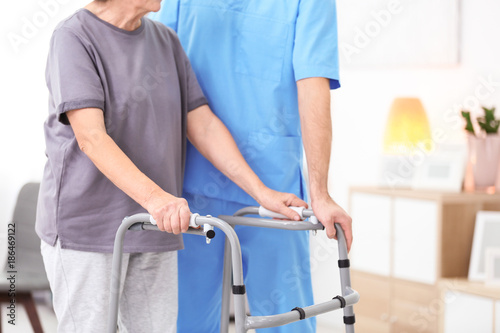 Senior woman walking with assistance of young caregiver indoors
