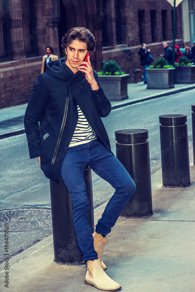 City life. Young handsome man wearing black fashionable trench coat with  hood, blue jeans, boots, sitting