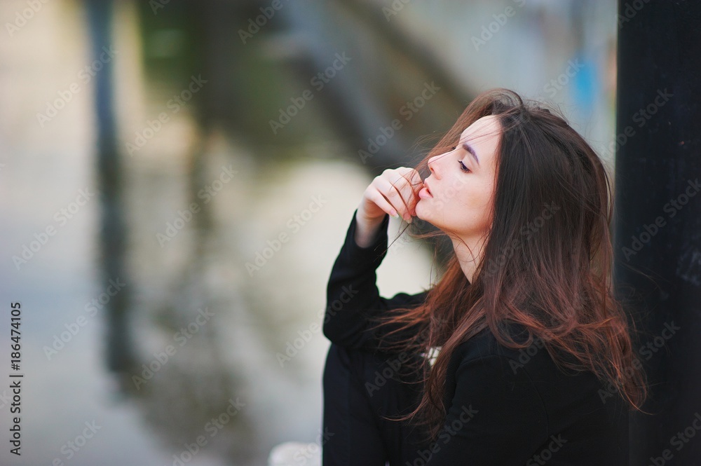 Portrait of a dreamy beautiful woman sitting in the street, shredded by wind long hair. Side view close-up on blurred background.