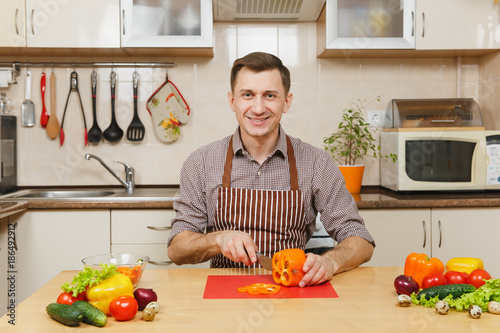 Bild auf Leinwand Handsome smiling caucasian young man in an apron, brown shirt sitting at table, cuts vegetable for salad with knife in light kitchen