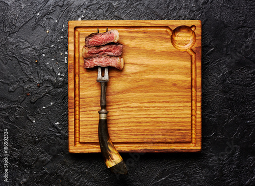 Slices of medium rare grilled Steak on meat fork on wooden meat cutting board. Black concrete background. Copy space. Top view.