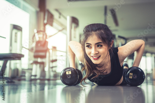 Asian young woman doing push ups with dumbbell on floor in fitness gym and equipment background. Workout and Sport Exercise concept. Healthy and Happiness concept. Beauty and Body build up theme. © Shutter2U