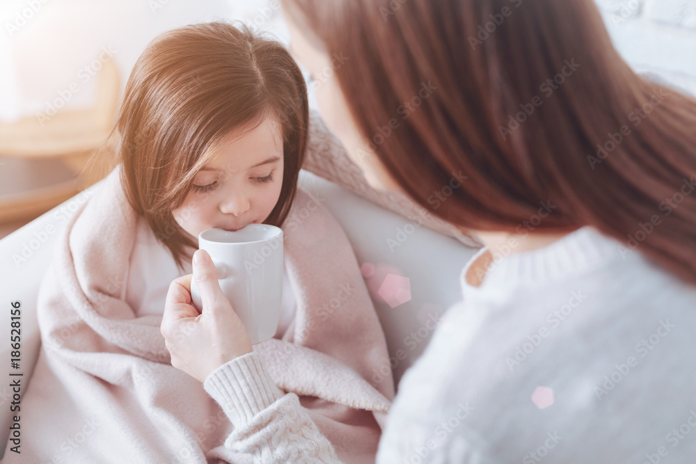 Tasty tea. Attractive brunette holding white cup in left hand and looking at her daughter while sitting in front of her Tasty tea. Attractive brunette holding white cup in left hand and looking at her daughter while sitting in front of her