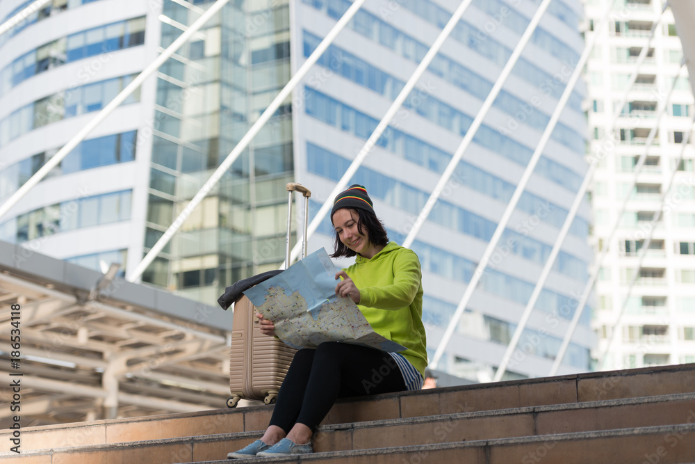 Happy female tourist with suitcase exploring map while sitting in city building, Travel concept