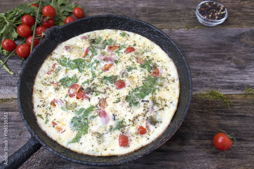 A beautiful omelette with greens and tomatoes in an old frying pan on a wooden background.